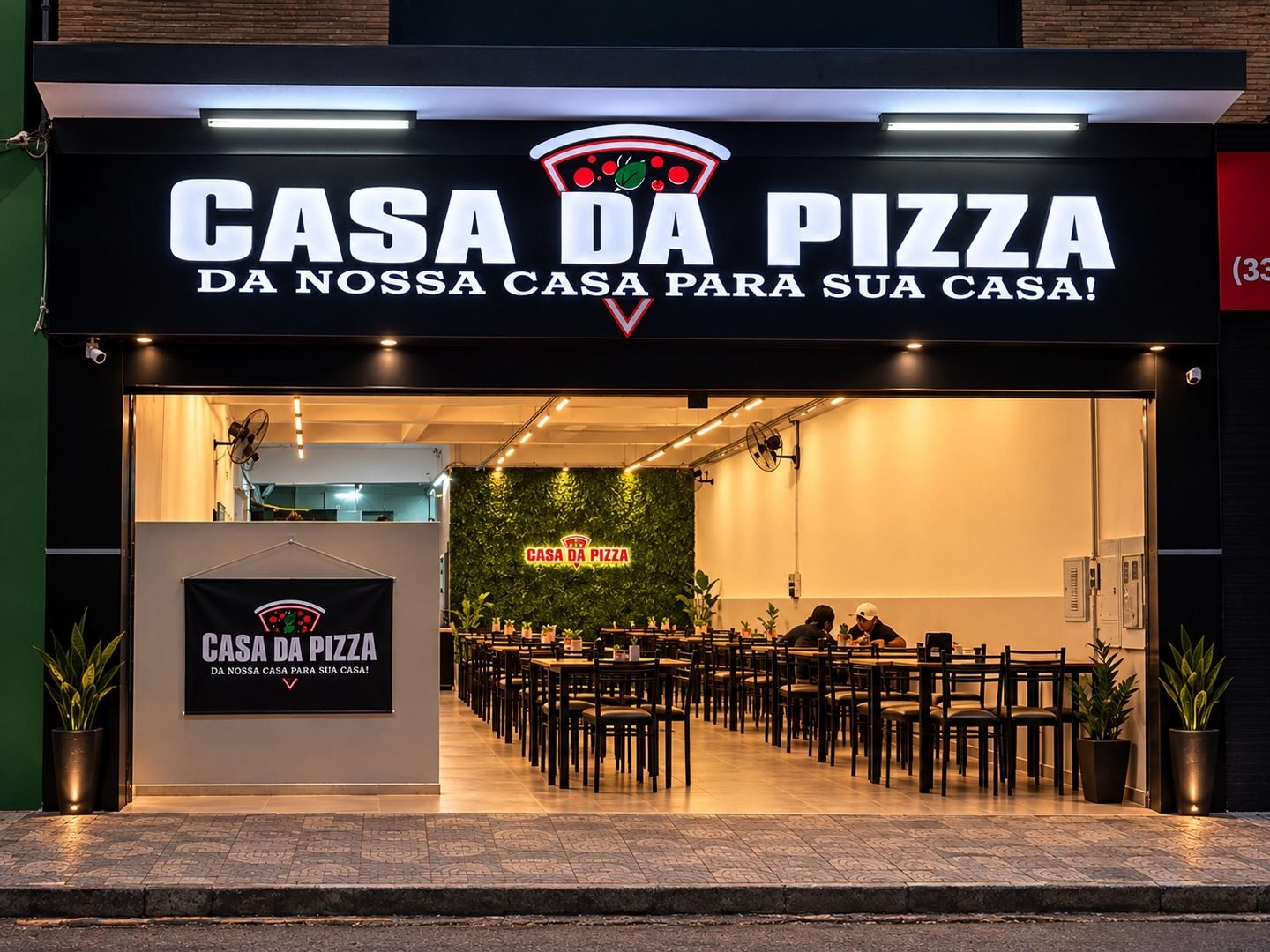 Fachada da Casa da Pizza à noite, com o letreiro aceso e o salão iluminado, com mesas e jardim vertical ao fundo.